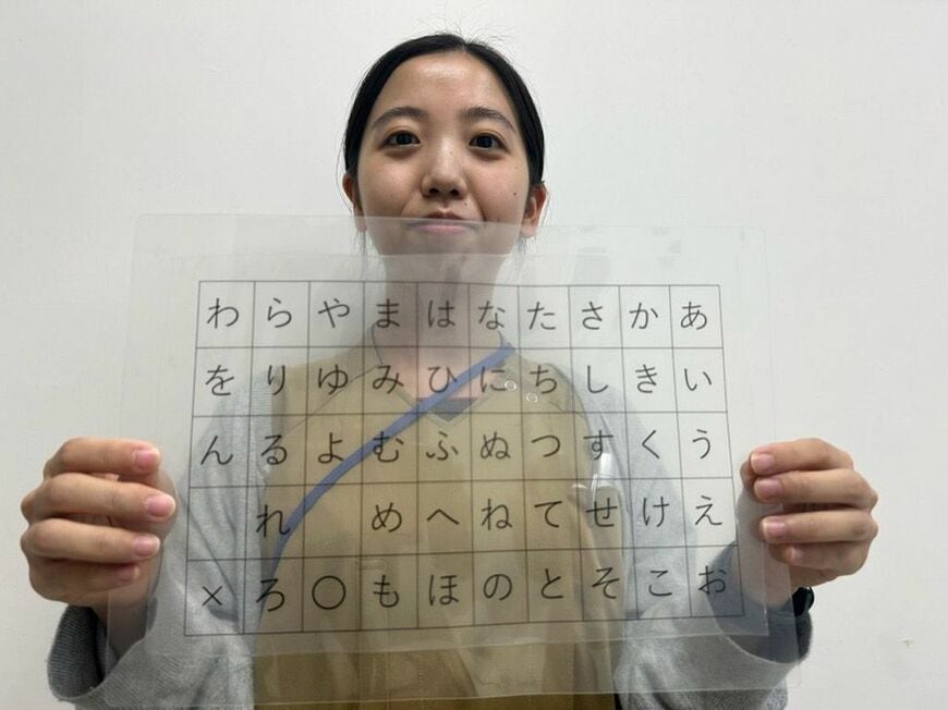 従来の意思疎通で使ってきた透明アクリル板の50音表。板越しに向き合い、一文字ずつ指しては反応を待つ。持っているのは言語聴覚士の屋宜氏