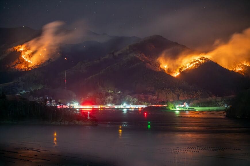 白煙を上げて山肌を焦がす炎。沿岸の民家付近の赤い光は消防車（写真：飯塚伸夫撮影）