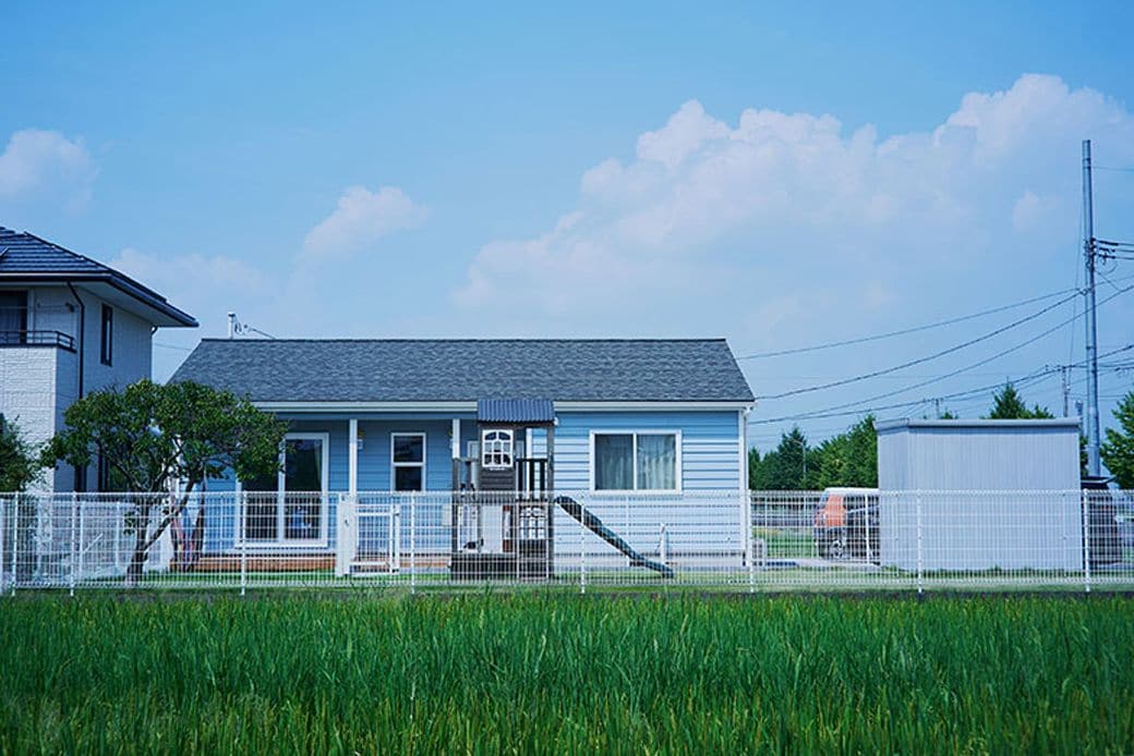 田んぼのグリーンにペールブルーの愛らしい外観が映えるみなこさんの家。伸びやかな周辺環境も含めアメリカンハウスの雰囲気そのもの（写真撮影：相馬ミナ）