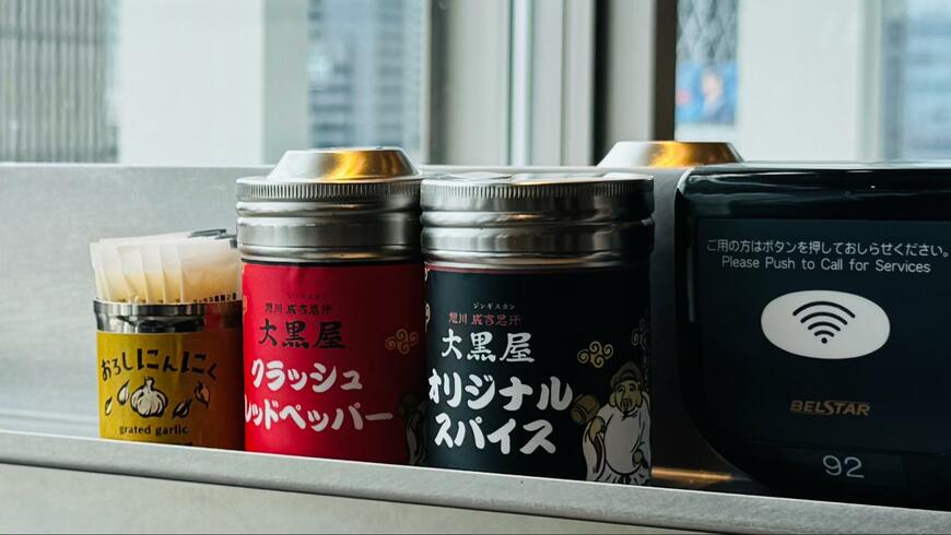 卓上スパイス棚。左から「おろしにんにく」「大黒屋クラッシュレッドペッパー」「大黒屋オリジナルスパイス」、呼び出しボタンが並ぶ（写真：筆者撮影）