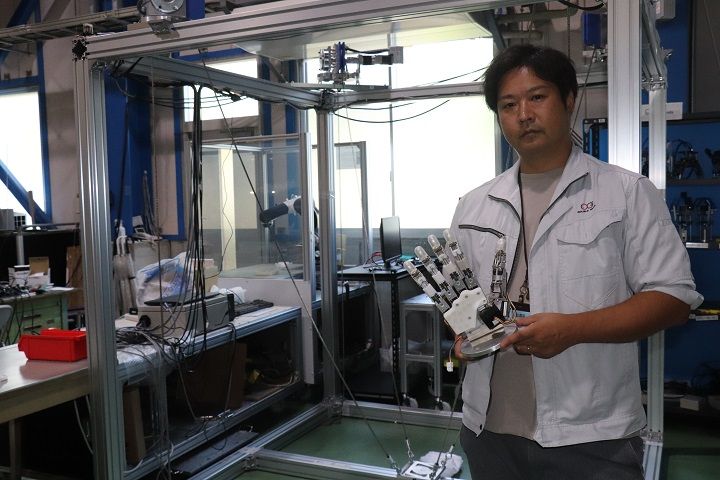 ゴクーに取り付けるロボットハンドを持つ和田始竜ダブル技研専務。後ろが遠隔就労システムの実験スペース（記者撮影）