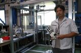 ゴクーに取り付けるロボットハンドを持つ和田始竜ダブル技研専務。後ろが遠隔就労システムの実験スペース（記者撮影）