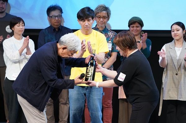 最優秀賞「うむさんラブ賞」を受賞した「看取り沖縄」プロジェクトの安里裕子さん（右）。沖縄国際映画祭実行委員長の大﨑洋氏から記念の泡盛が贈られた（写真：吉本興業提供）